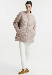 Gequilted beige jas met ronde halslijn, drukknoopjes en zijzakken, gecombineerd met witte broeken en bruine suède schoenen. Gladde textuur.