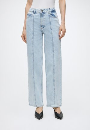 Jean taille haute bleu clair à jambes larges avec des coutures sur le devant, porté avec des chaussures à talons noires à bout pointu.