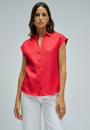 Femme portant une blouse sans manches rouge vif boutonnée et un pantalon blanc, debout devant un fond gris clair uni.