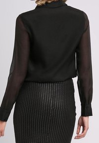 Blusa nera con maniche lunghe trasparenti e colletto classico, abbinata a una gonna a matita nera con texture e motivo a griglia.