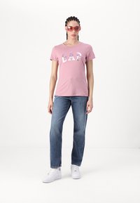 Rožinė medvilninė marškinėliai su „GAP“ logotipu raštuotame dizaine, suporuoti su mėlynais tiesiais džinsais ir baltais sportbačiais. Modelis nešioja rožinius akinius nuo saulės.