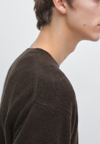 Brauner Strickpullover mit rundem Ausschnitt. Der Stoff hat eine weiche, strukturierte Oberfläche und sichtbare Nähte an den Schultern.