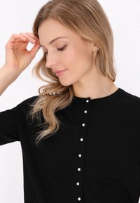 Maglione nero a maglia con scollo rotondo e sette bottoni decorativi in perla lungo il davanti. Tessuto liscio e design aderente.