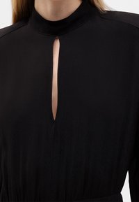 Schwarzes Kleid mit hohem Kragen und einem Schlitzöffnungsbereich am Hals. Glatte Stofftextur mit subtiler Glanz. Figurbetontes Design mit gerafftem Taillenbereich.