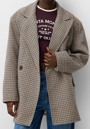Personne portant un manteau à carreaux beige et marron sur un t-shirt graphique bordeaux raccourci et un jean bleu, tenant le revers du manteau.