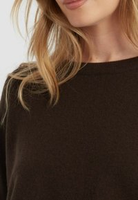 Maglione a maglia marrone con scollatura rotonda, texture morbida e dettagli di cucitura discreti sul colletto. Capelli parzialmente visibili di lato.