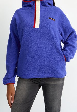 Personne portant un pull polaire bleu avec une patte de bouton-pression rouge et blanche et un logo Columbia, associé à un jean gris foncé.