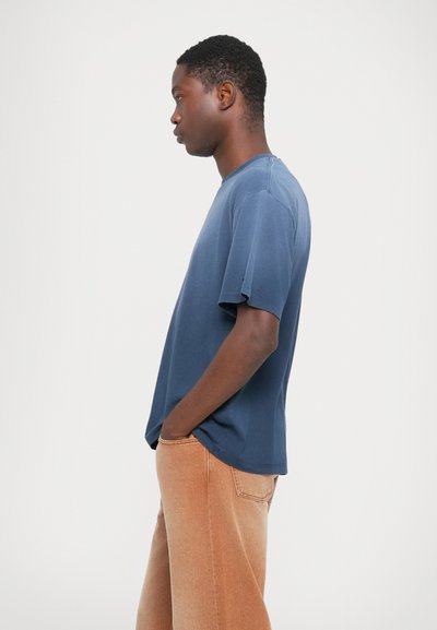 Modra majica s krojem okroglega vratu iz bombaža s kratkimi rokavi, ki ima ohlapen kroj in rahlo teksturirano površino, v kombinaciji s bež širokimi hlačami.