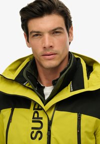 Yellow and black jacket with zippers, large "SUPER" logo, and a contrasting inner layer. Fabric appears smooth and weather-resistant. Black accents on shoulders.