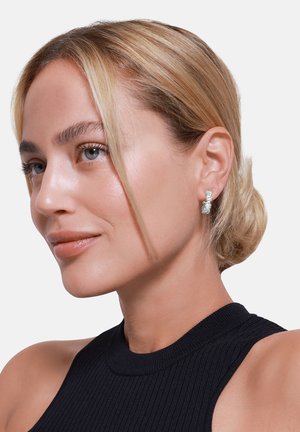 Silberne Ohrstecker mit strukturiertem Design und einem kleinen Steinakzent, präsentiert auf einem hellen Hintergrund. Modell trägt ein schwarzes Rippenshirt.