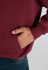 Mano appoggiata all'interno della tasca frontale di un felpa color bordeaux, con dettagli visibili del polsino, e parte di jeans in denim blu su uno sfondo semplice.