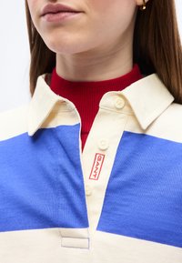 Femme portant un polo à rayures bleues et crème superposé à un col roulé rouge, montrant les détails du col et une petite étiquette logo.
