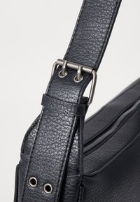 Borsa a tracolla in pelle nera con superficie testurizzata, accessori in metallo argento e anelli metallici per la regolazione. Scomparto con zip visibile.