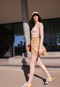 Top cropped às riscas, saia mini floral com barra em babados, sapatilhas brancas com detalhes em laranja e preto, e um chapéu tipo balde bege.
