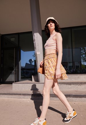 Top cropped às riscas, saia mini floral com barra em babados, sapatilhas brancas com detalhes em laranja e preto, e um chapéu tipo balde bege.