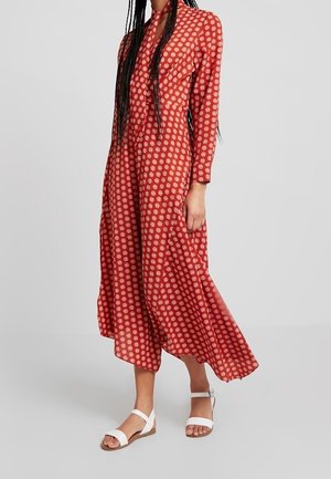Femme portant une robe midi rouge fluide avec des motifs circulaires blancs et des manches longues, associée à des sandales blanches à bouts ouverts.