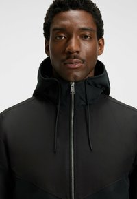 Chaqueta negra con cremallera alta, que presenta una textura suave y un diseño bicolor. Destacan la cremallera plateada y los acentos de cordón.