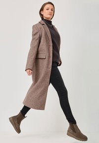 Brown wool blend coat with a tailored design, worn over a dark sweater and slim-fit black pants. Features ankle-height brown suede boots.