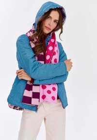 Tyrkysová prešívaná páperová bunda s kapucňou, zdobená kvetinovými vzormi. Doplnok tvorí ružovo-fialový pruhovaný šál s bodkami. Zavesené ruky prekrížené.