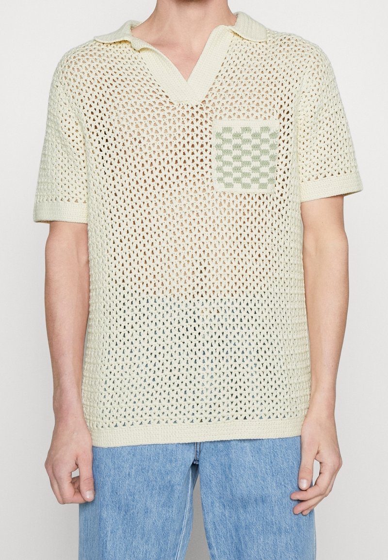 Homme portant un polo en crochet crème à manches courtes avec un col large et une poche à motif à carreaux, associé à un jean bleu clair.