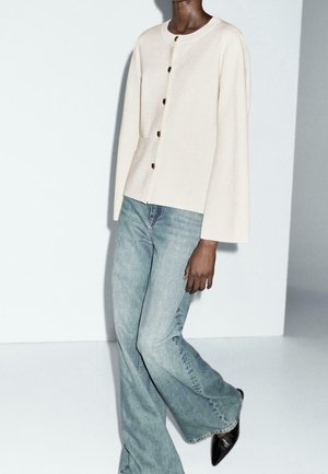 Persoon die een crèmekleurig vest met knopen draagt, lichtblauwe wijde jeans en zwarte puntige schoenen, staand tegen een effen lichte achtergrond.