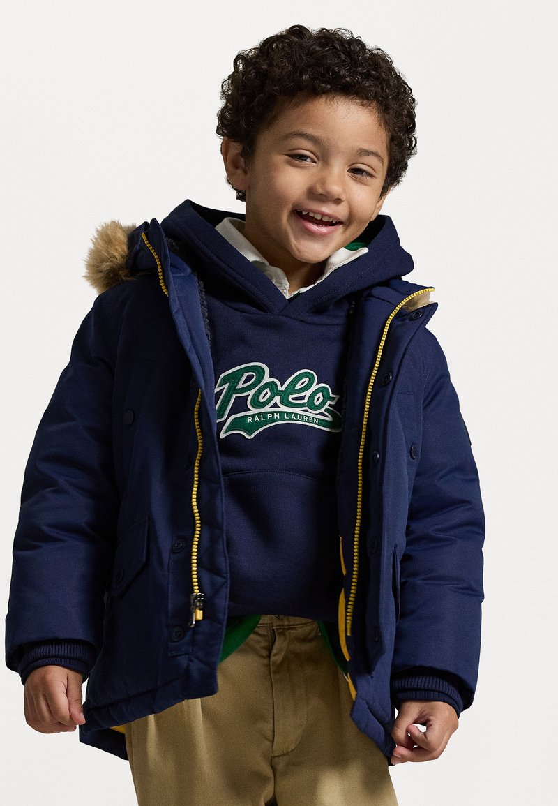 Σκούρο μπλε επενδυμένο μπουφάν με κίτρινο φερμουάρ και κουκούλα με γούνινη επένδυση, φορεμένο πάνω από μια σκούρα φούτερ με το λογότυπο "Polo", συνδυασμένο με μπεζ παντελόνι.