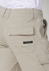 Pantaloni cargo bej cu talie elastică, multiple buzunare, țesătură ușoară, detalii cusute și o etichetă neagră pe spate.