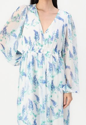 Kobieta nosząca białą sukienkę z długim rękawem w niebiesko-zielony wzór kwiatowy i ze szczegółem marszczonej talii.