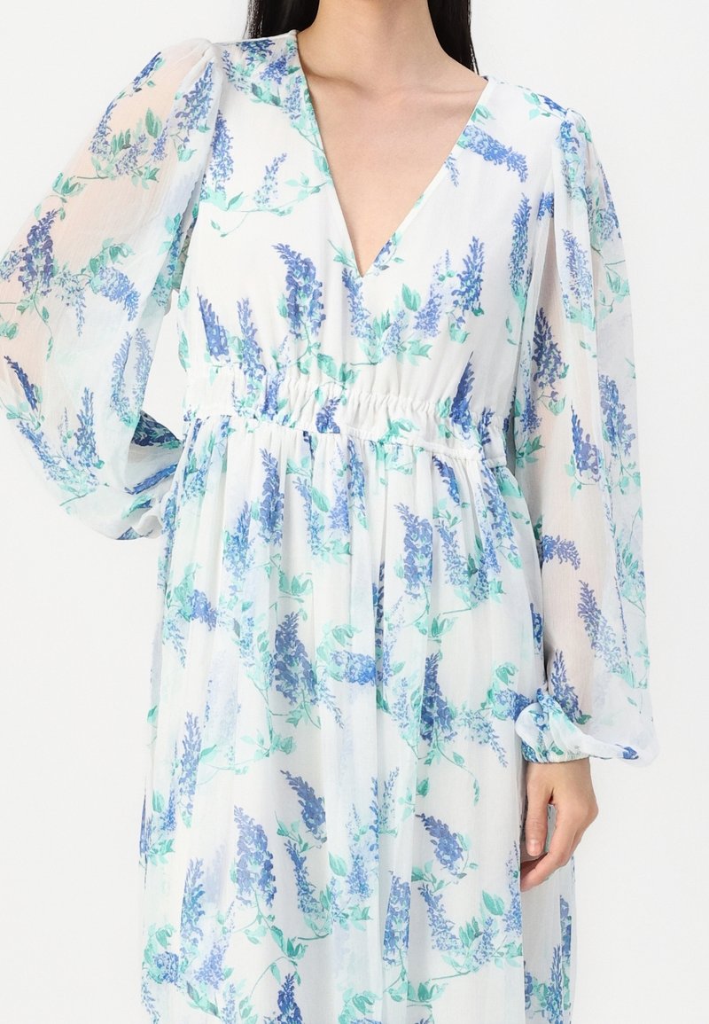 Femme portant une robe blanche à manches longues avec un motif floral bleu et vert et un détail froncé à la taille.