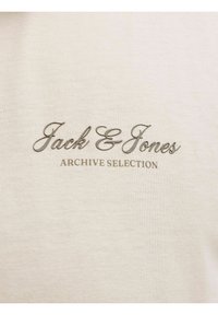 Μπεζ ύφασμα με την επιγραφή 'Jack & Jones' με γραφή υπογραφής και 'ARCHIVE SELECTION' με κεφαλαία γράμματα τυπωμένα στο κέντρο της επιφάνειας.