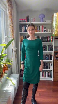 Vrouw in groene jurk en zwarte laarzen staat in een kamer met een boekenkast, een plant bij het raam en een houten vloer.