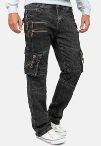 Pantaloni cargo in denim nero con molte tasche con cerniera, tessuto strutturato, dettagli trapuntati e una vestibilità affusolata, abbinati a sneaker bianche.