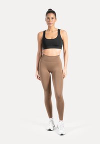 Brassière de sport noire associée à un legging marron taille haute, les deux réalisés dans un matériau doux et extensible. Modèle portant des baskets blanches.