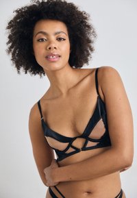 Svart bralette med gjennomsiktige paneler, blonderdetaljer og justerbare stropper. Designet inkluderer utskjæringer og hardware i gulltone.