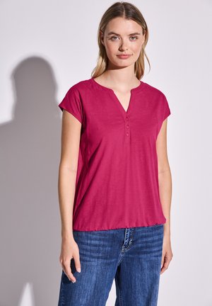 Magenta kortärmad topp med V-ringning och tre-knappsstängning, tillsammans med blåjeans. Slät textur. Enkelt mönster, inga mönster.