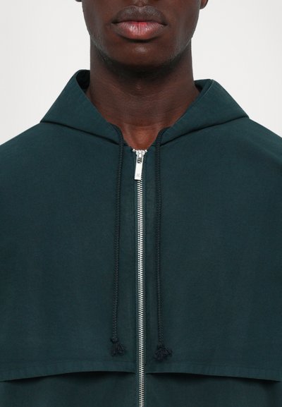 Κοντινό πλάνο ενός ατόμου που φοράει μια σκούρα πράσινη μπουφάν με χνουδωτή κουκούλα, με ασημί φερμουάρ μπροστά και κορδόνια προσαρμογής.