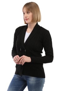 Schwarzer Strickcardigan mit Schalkragen, doppelter Knopfverschluss und gerippten Bündchen. Über einem weißen T-Shirt getragen und mit blauen Jeans kombiniert.