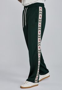 Pantalones deportivos verde oscuro con una franja lateral floral en blanco y rojo, cinturilla elástica y puños acanalados, que cuentan con bolsillos delanteros y bordado del logo.