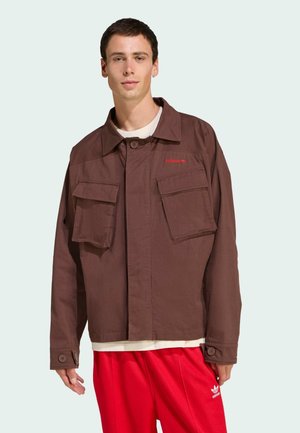 Brown cotton jacket with two front pockets, button closure, and a collar. Features red logo and buttoned cuffs. Worn over a white shirt.