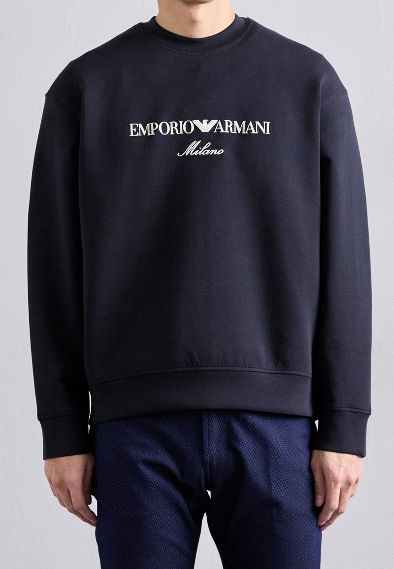 Námořnická mikina s kulatým výstřihem, která má velký bílý nápis "Emporio Armani Milano" přes hrudník, vyrobena z bavlněného materiálu.