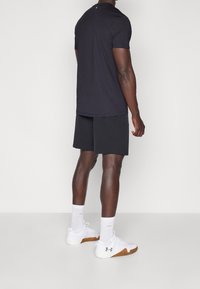Schwarzes Sportshirt mit kurzen Ärmeln, kombiniert mit schwarzen Shorts, weißen Sportsocken und weißen Schuhen mit braunen Sohlen.