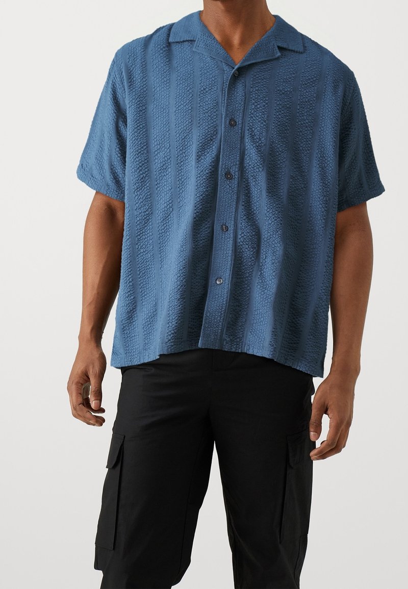 Homme portant une chemise bleue texturée à manches courtes boutonnée et un pantalon cargo noir sur un fond uni.