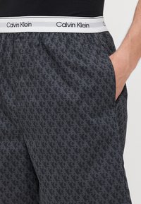Sötét szürke Calvin Klein rövidnadrág, amelyen logós minta található. Rugalmas derékpánttal rendelkezik, logós részletekkel és oldalsó zsebekkel.