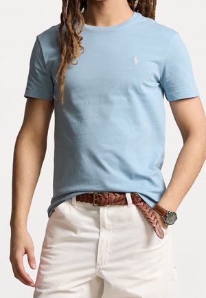 Mężczyzna w jasnoniebieskim t-shircie z krótkim rękawem, białych spodniach z brązowym plecionym skórzanym paskiem oraz brązowym zegarkiem na lewej ręce na białym tle.