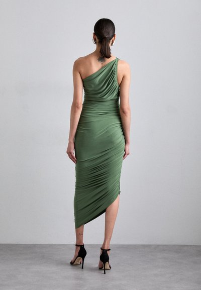 Asymmetrical green dress made of smooth, draped fabric, featuring a gathered design on one shoulder and a thigh-high slit, paired with black heels.