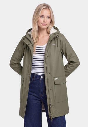 Blonde Frau trägt eine olivgrüne Kapuzen-Regenjacke über einem gestreiften Shirt und blauen Jeans, steht mit den Händen hinter dem Rücken.
