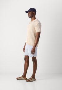 Beige knitted polo shirt with a collar, light blue textured shorts, and brown slide sandals. Model wearing a navy cap.