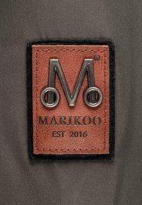 Etiqueta de cuero con el logotipo "M" en relieve, contorneado con hardware plateado. El texto dice "MARIKOO EST 2016" sobre un fondo de tela gris.