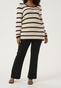 Pull en maille blanche avec des rayures noires, manches longues et encolure ronde, associé à un pantalon noir. La texture est en maille ajourée et ample.