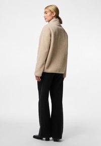 Jachetă din fleece bej cu guler înalt, croială lejeră și textură moale, asociată cu pantaloni negri cu croi larg și cizme până la gleznă.